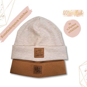 Könnte beinhalten: Eine beige Mütze mit einem braunen Kunstleder-Patch, auf dem "Wild One" steht, und einem weiteren Patch, auf dem "So Free & Wild + Wanderlust" steht. Die Mütze ist umdrehbar und hat eine braune Seite.