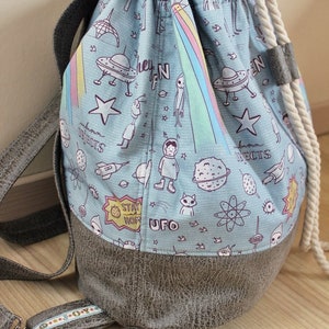 Puede incluir: Una mochila azul con cord&oacute;n y asa de cuerda blanca. La mochila est&aacute; hecha de tela con fondo azul claro y un estampado de tem&aacute;tica extraterrestre blanco, negro y rosa. El texto "Stay Home" y "UFO" est&aacute; impreso en la mochila. La mochila tiene un fondo de cuero marr&oacute;n.