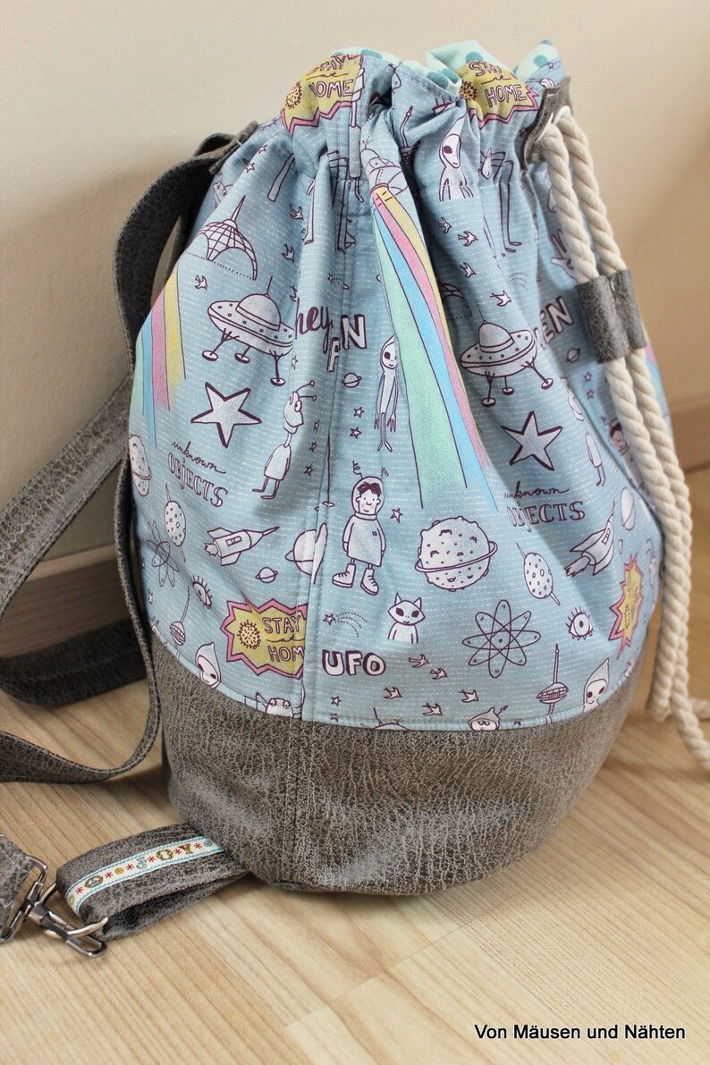 Puede incluir: Una mochila azul con cord&oacute;n y asa de cuerda blanca. La mochila est&aacute; hecha de tela con fondo azul claro y un estampado de tem&aacute;tica extraterrestre blanco, negro y rosa. El texto "Stay Home" y "UFO" est&aacute; impreso en la mochila. La mochila tiene un fondo de cuero marr&oacute;n.