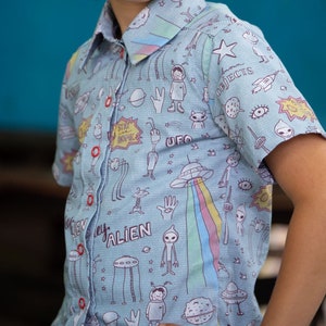 Puede incluir: Una camisa de manga corta azul claro con botones y un estampado colorido de extraterrestres y OVNIs. La camisa tiene un cuello blanco y botones. El estampado incluye las palabras "Stay Home", "Alien" y "UFO".