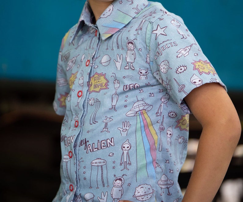 Puede incluir: Una camisa de manga corta azul claro con botones y un estampado colorido de extraterrestres y OVNIs. La camisa tiene un cuello blanco y botones. El estampado incluye las palabras "Stay Home", "Alien" y "UFO".
