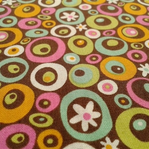 Könnte beinhalten: Ein brauner Stoff mit einem sich wiederholenden Muster aus bunten Kreisen und stilisierten Blumen. Die Kreise sind in den Farben Rosa, Grün, Gelb und Weiß gehalten. Die Blumen sind weiß mit rosa Zentren.