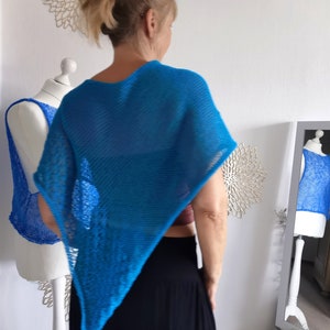 Könnte beinhalten: Ein leuchtend blauer, offen gewebter Poncho. Das dreieckige Kleidungsstück ist über einer Person drapiert und zeigt sein luftiges Design. Der Poncho ist ein helles, einfarbiges Blau und scheint handgefertigt zu sein. Ein passendes Oberteil ist auf einer Schaufenquin zu sehen.