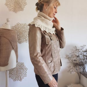 Könnte beinhalten: Eine Frau trägt eine hellbraune Lederjacke und einen cremefarbenen Strickkragen mit gewelltem Rand. Die Jacke hat Taschen und einen Schnallenverschluss. Eine Schaufensterpuppe zeigt ein Strickkleidungsstück. Dekorative Wandkunst ist im Hintergrund zu sehen.