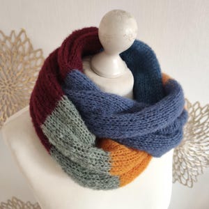 Könnte beinhalten: Ein grob gestrickter Infinity-Schal mit Color-Block-Design in den Farben tiefes Burgunderrot, gedämpftes Salbeigrün, leuchtendes Senfgelb und Staubblau. Der Schal ist zu einer kreisförmigen Schlaufe gewickelt und zeigt ein weiches, flauschiges Garn.