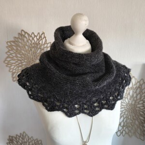 Könnte beinhalten: Ein dunkel anthrazitfarbener Strickkragen mit hohem Hals und einem dekorativen, gehäkelten Wellenrand. Das Accessoire weist ein durchgehendes, strukturiertes Strickmuster auf und wird auf einer weißen Schaufensterpuppe mit einer silbernen Kette präsentiert.