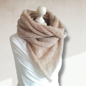 Könnte beinhalten: Ein hellbeige, dreieckiger Schal ist um eine weiße Schaufensterpuppe drapiert. Der Schal besteht aus einem weichen, gestrickten Material. Der Schal ist um den Hals und die Schultern der Schaufensterpuppe gewickelt und erzeugt einen gemütlichen und stilvollen Look. Der Hintergrund ist hellbeige.
