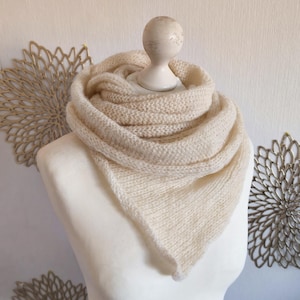 Könnte beinhalten: Ein cremefarbener, dreieckiger Strickschal mit einer weichen, flauschigen Textur. Der Schal ist in mehreren Lagen um den Halsbereich drapiert und weist ein durchgehendes glatt rechts gestricktes Muster mit einer spitz zulaufenden Vorderkante auf.