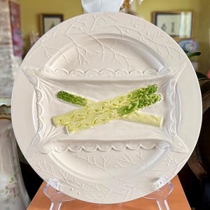 May include: A white ceramic serving plate with a decorative asparagus design. The plate is displayed on a stack of books with titles including "French by Design" and "Adventures with Old Houses".