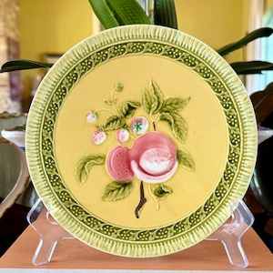 May include: A round ceramic plate with a pale yellow center and a textured green border. The center features a raised, three-dimensional relief of a pink peach with green leaves and small white flowers. The plate is displayed on a clear stand.
