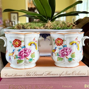 May include: Two white porcelain planters with floral and butterfly designs, sitting atop a stack of books. The planters have small handles and are filled with moss. A decorative plate and pitcher are also visible.
