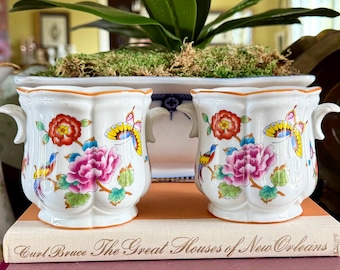 May include: Two white porcelain planters with floral and butterfly designs, sitting atop a stack of books. The planters have small handles and are filled with moss. A decorative plate and pitcher are also visible.