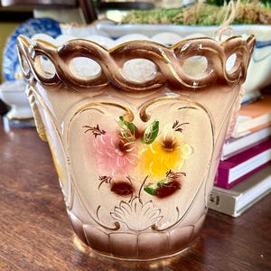 May include: A ceramic planter featuring a cream-colored body with a brown gradient rim and base. The exterior is decorated with a hand-painted floral motif in pink and yellow. The top edge has a decorative openwork lattice design.