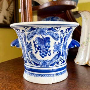 May include: A white ceramic planter with a blue transferware pattern featuring grape clusters, leaves, and ornate scrollwork. The vessel has a flared rim, a tapered body, and two small blue handles on opposite sides, finished with a decorative border.