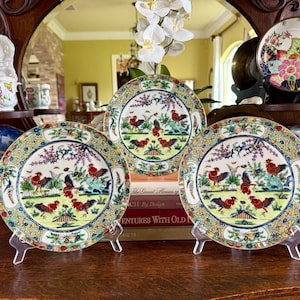 May include: Three decorative ceramic plates with rooster and floral designs. The plates are displayed on clear stands, showcasing a detailed landscape scene with roosters, trees, and butterflies. The plates have a colorful border.