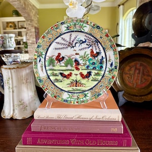 May include: A decorative plate with a rooster design, resting on a stack of books. The plate features a colorful border and a central scene with roosters and other elements. The books have titles like "French by Design."