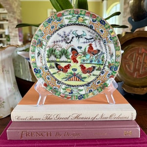 May include: A decorative porcelain plate with a colorful rooster design. The plate features a detailed border and is displayed on a clear stand, resting on a stack of books. The plate's design includes roosters, birds, and floral elements.