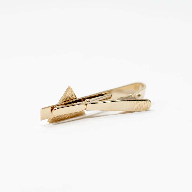 Puede incluir: Un clip de corbata dorado con un dise&ntilde;o geom&eacute;trico. El clip presenta una base rectangular y un acento triangular. El clip est&aacute; dise&ntilde;ado para asegurar una corbata y est&aacute; hecho de un material met&aacute;lico brillante.