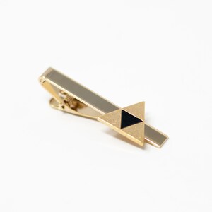 Puede incluir: Un pasador de corbata dorado con un acabado texturizado. El pasador presenta un dise&ntilde;o triangular con un tri&aacute;ngulo negro en el centro. El pasador de corbata est&aacute; dise&ntilde;ado para asegurar una corbata.