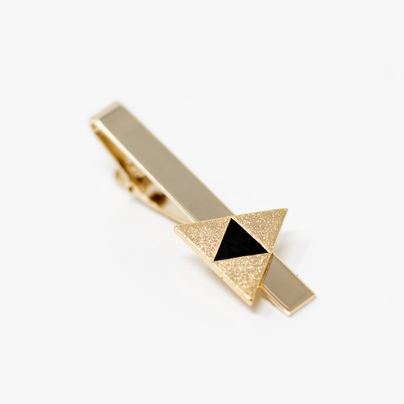 Puede incluir: Un pasador de corbata dorado con un dise&ntilde;o triangular. El tri&aacute;ngulo presenta un exterior dorado texturizado y brillante y un tri&aacute;ngulo negro en el centro. El pasador tiene forma rectangular y un cierre.