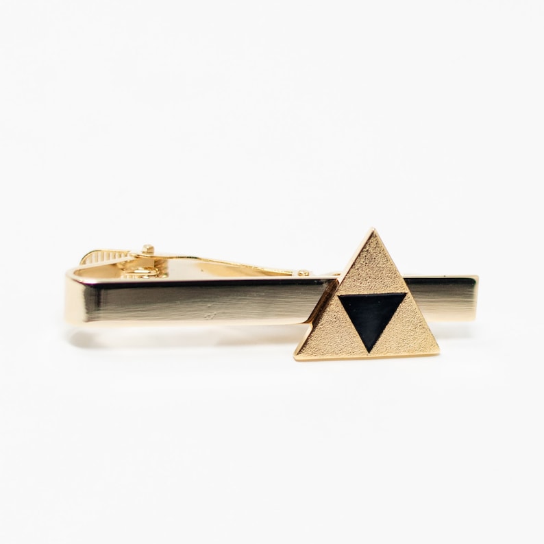Puede incluir: Pinza de corbata dorada con un emblema triangular con un tri&aacute;ngulo negro. El clip de corbata tiene un dise&ntilde;o cl&aacute;sico y un acabado brillante. Un accesorio elegante para ropa formal.