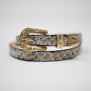 May include: A gray snakeskin belt with a gold-toned buckle. The buckle has a detailed design and a pointed tip.