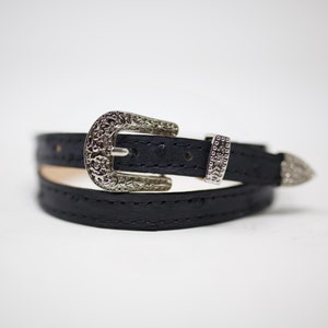 May include: A black leather belt with a silver ornate buckle and a silver tip. The belt is folded in half and the buckle is closed.