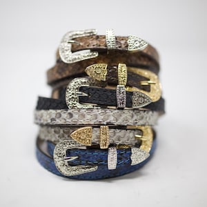 May include: A stack of five different colored leather belts with silver and gold buckles. The belts are all different colors and patterns, including brown, black, white, and blue.