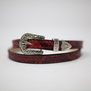 May include: A red snakeskin belt with a silver ornate buckle. The buckle has a decorative design and a pointed tip.