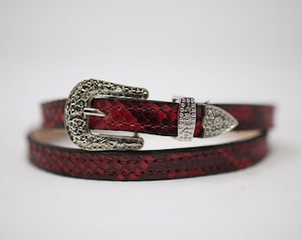 Puede incluir: Un cinturón de piel de serpiente roja con una hebilla plateada adornada. La hebilla tiene un diseño decorativo y una punta puntiaguda.