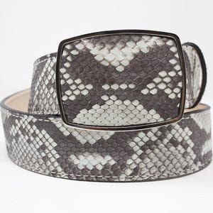 May include: A coiled belt with a snakeskin pattern in shades of gray and white. The belt features a rectangular buckle with a silver-colored frame. The design is a neutral color palette, suitable for various outfits.