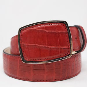 May include: A red leather belt with a textured, crocodile-like pattern. The belt features a large, rectangular silver buckle. The belt is rolled up, showcasing the texture and color. The belt is a fashion accessory.
