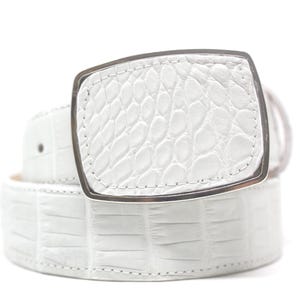 May include: A white belt with a textured, crocodile-like pattern. The belt features a rectangular silver buckle. The belt is rolled up, showcasing the texture and design.