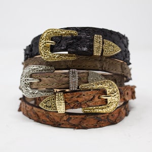 May include: Three leather belts with ornate metal buckles. The belts are stacked on top of each other, with the black belt on top, followed by a brown belt, and then a tan belt.