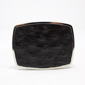 May include: A black ostrich leather belt buckle with a silver-toned metal frame.