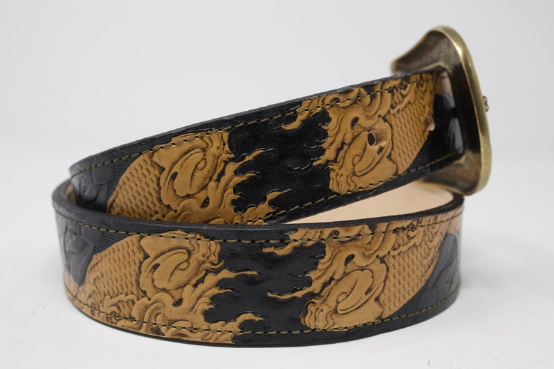 May include: A brown leather belt with a black and brown embossed design. The belt has a large, ornate brass buckle.