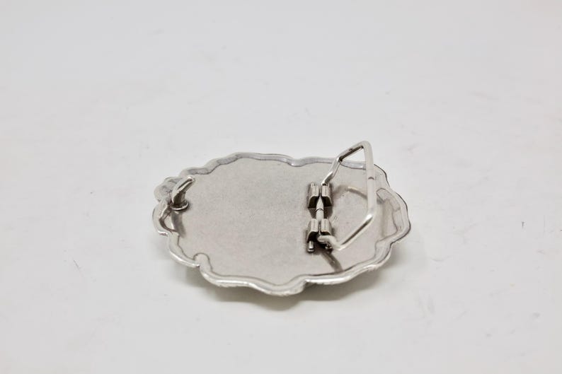 Puede incluir: Hebilla de cintur&oacute;n de metal plateado con forma ovalada y borde festoneado decorativo. La parte posterior de la hebilla presenta un mecanismo de barra y pasador para sujetar a un cintur&oacute;n. La hebilla est&aacute; dise&ntilde;ada para ser duradera y funcional.