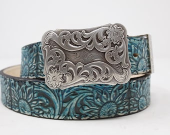 Peut inclure: Une ceinture en cuir turquoise avec une boucle argentée. La ceinture présente un motif floral et une boucle décorative au design orné. La ceinture est enroulée, mettant en valeur le savoir-faire détaillé. La ceinture mesure environ 100 cm.