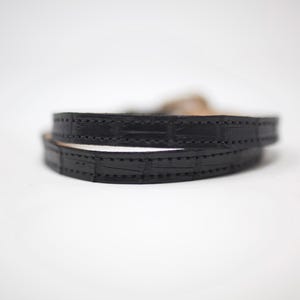 Puede incluir: Un brazalete de cuero negro con un patr&oacute;n de cocodrilo y costuras.