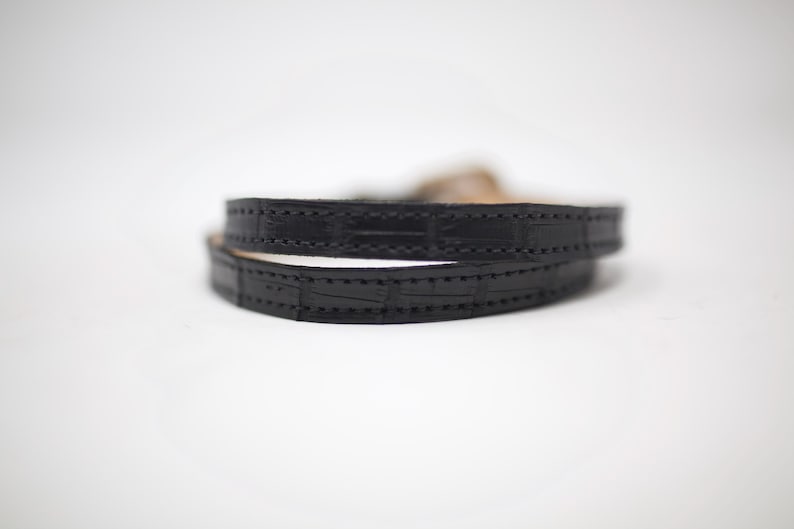 Puede incluir: Un brazalete de cuero negro con un patr&oacute;n de cocodrilo y costuras.