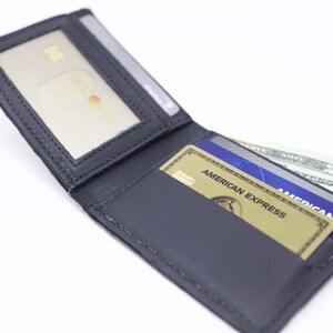 Peut inclure: Un portefeuille en cuir noir avec une fen&ecirc;tre d'identit&eacute; transparente, ouvert pour r&eacute;v&eacute;ler deux cartes de cr&eacute;dit et un billet de vingt euros. Les cartes de cr&eacute;dit sont une carte American Express et une carte bleue avec le texte "AMERICAN EXPRESS" dessus.