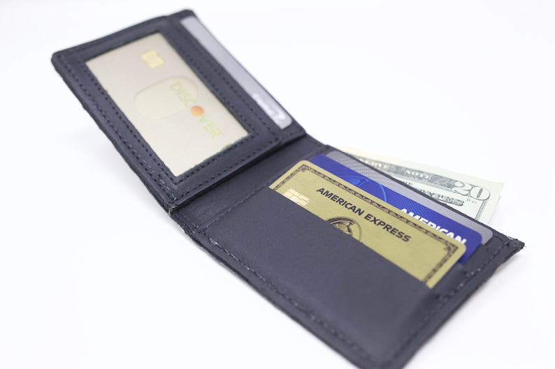 Peut inclure: Un portefeuille en cuir noir avec une fen&ecirc;tre d'identit&eacute; transparente, ouvert pour r&eacute;v&eacute;ler deux cartes de cr&eacute;dit et un billet de vingt euros. Les cartes de cr&eacute;dit sont une carte American Express et une carte bleue avec le texte "AMERICAN EXPRESS" dessus.