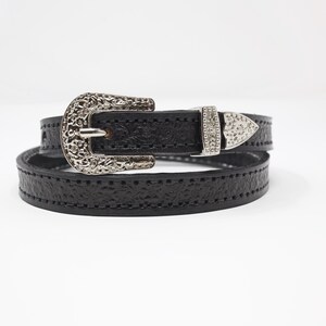 May include: Black leather belt with a decorative silver buckle and a matching silver tip. The belt is wrapped around itself, showcasing the textured leather and the intricate details of the buckle design.