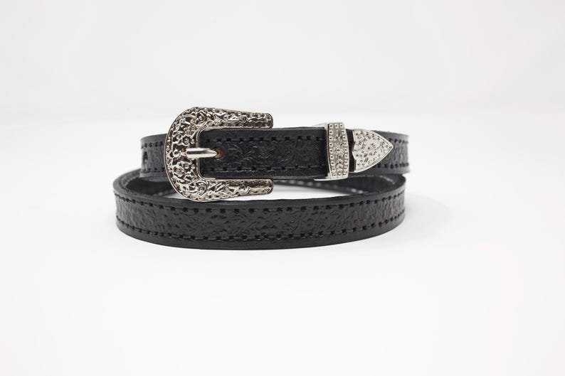 May include: Black leather belt with a decorative silver buckle and a matching silver tip. The belt is wrapped around itself, showcasing the textured leather and the intricate details of the buckle design.