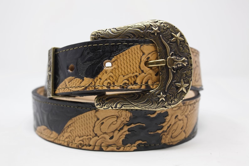 May include: A black and tan leather belt with an ornate brass buckle featuring a star design. The belt is decorated with an embossed pattern of fish and waves.