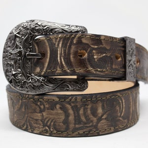 May include: A brown leather belt with an intricate silver buckle. The belt has a floral pattern embossed into the leather.