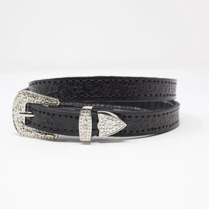 May include: Black leather belt with a textured surface and silver-colored metal buckle. The buckle has a decorative design. The belt is wrapped around itself.