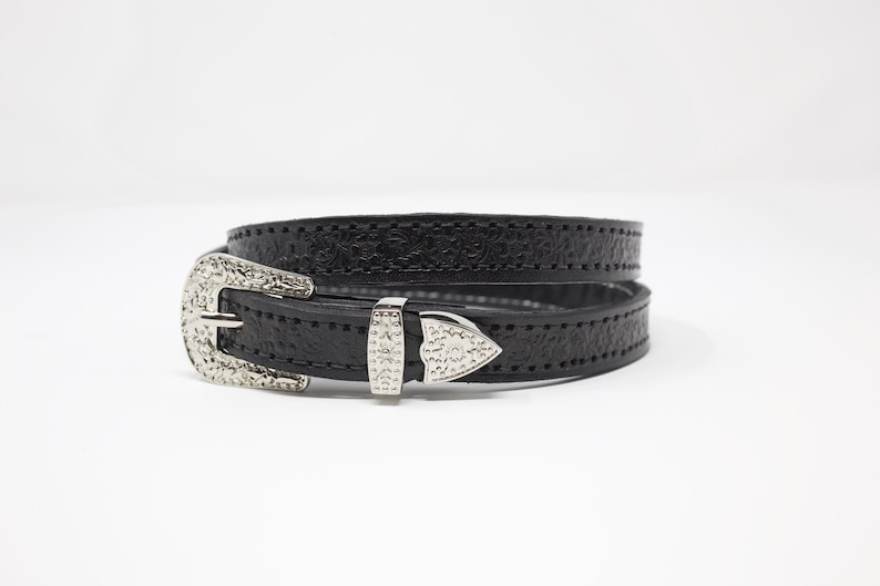 May include: Black leather belt with a textured surface and silver-colored metal buckle. The buckle has a decorative design. The belt is wrapped around itself.