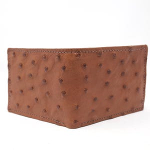 May include: A brown leather wallet with a distinctive ostrich skin texture. The wallet is rectangular and features a stitched border. The leather has a pattern of small, dark brown bumps. The wallet is set against a white background.