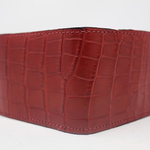 May include: A bifold wallet in a deep red color featuring a textured crocodile skin pattern. The wallet has visible stitching along the edges and a rectangular shape, presented against a plain, neutral background with a smooth finish.